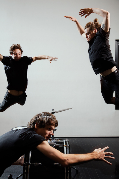 Two men jump into the air while their drummer hovers near his set